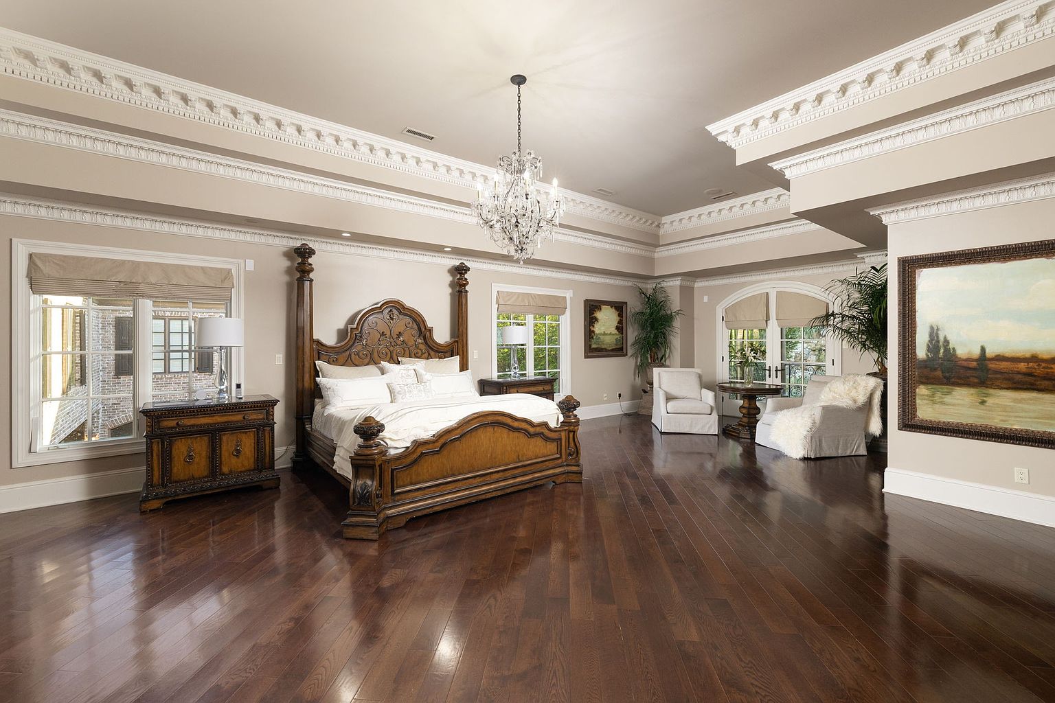 This is a luxurious primary bedroom featuring a four-poster bed with ornate wood carvings and plush white bedding. The room is spacious with dark hardwood floors, elegant crown molding, and a crystal chandelier. Large windows provide natural light, and there are seating areas with comfortable armchairs, creating a serene and inviting atmosphere.