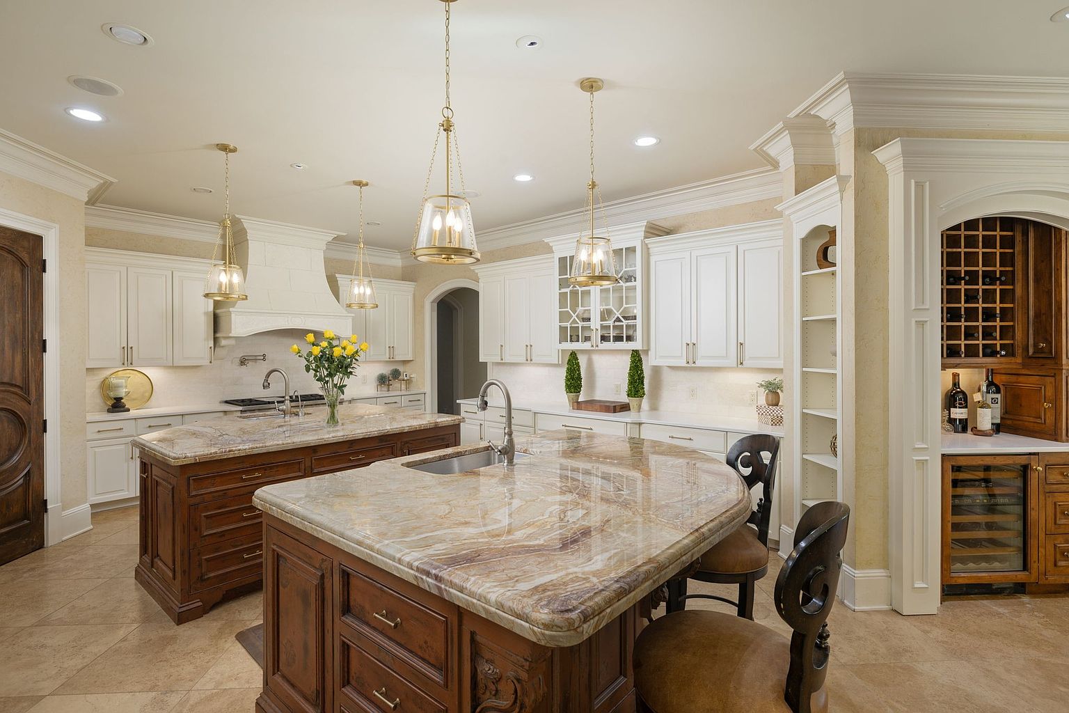 This is a well-lit, luxurious kitchen featuring two islands with marble countertops and dark wood cabinetry. The kitchen is equipped with white cabinets, pendant lighting, and high-end appliances. A wine cellar is visible in the background, adding to the upscale feel of the space.