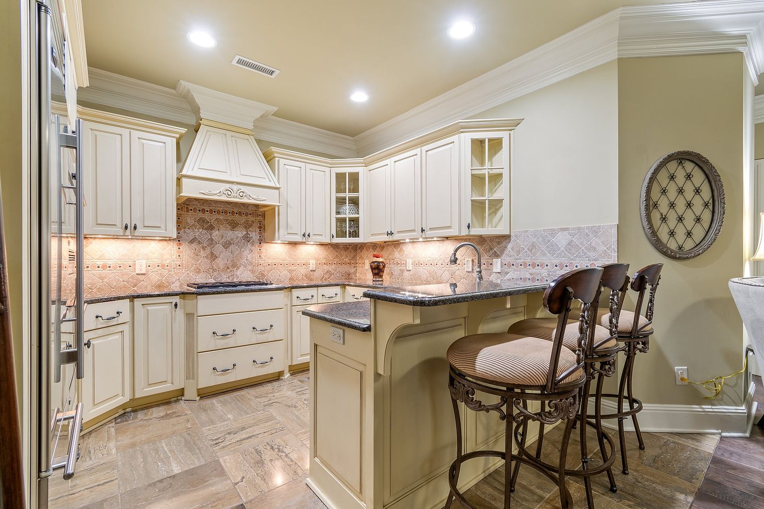 This is a well-lit kitchen featuring cream-colored cabinetry, granite countertops, and a decorative tile backsplash. The kitchen includes a central island with bar seating and stainless steel appliances. The flooring is a light-colored tile, and the overall style is traditional and elegant.
