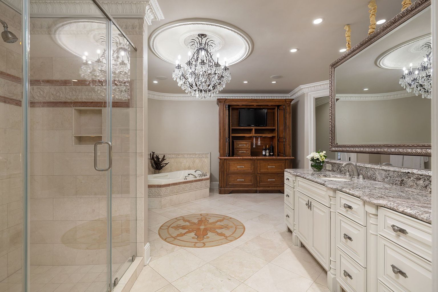 This is a luxurious primary bathroom featuring a glass-enclosed shower, a soaking tub, and a vanity with granite countertops. A large mirror hangs above the vanity, reflecting the elegant chandelier that illuminates the room. The flooring is tiled with a decorative medallion, and a wooden cabinet provides additional storage.