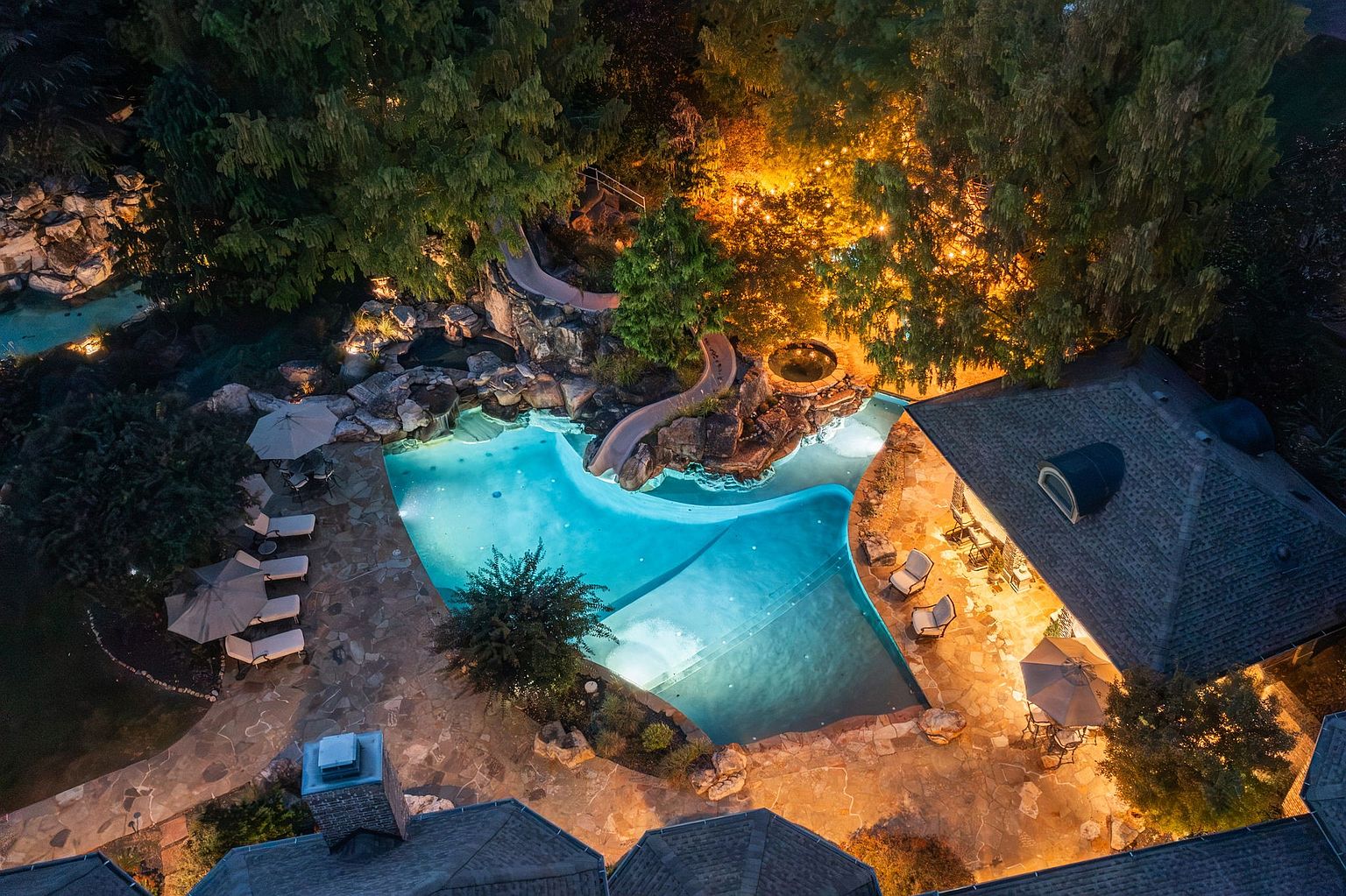 This aerial view showcases a luxurious backyard oasis featuring a uniquely shaped pool with integrated spa and a water slide built into a rock formation. The surrounding landscape includes mature trees, stone pathways, and outdoor seating areas, creating a private and inviting retreat. The property is well-lit, highlighting the architectural details and enhancing the overall ambiance.