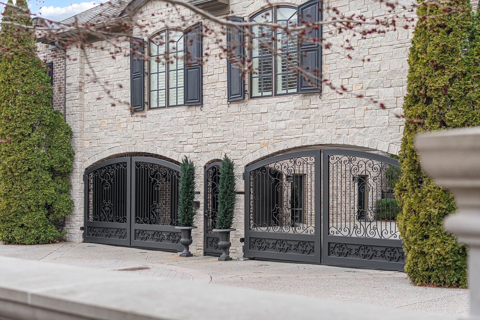 This is a front exterior view of a luxurious stone house featuring arched wrought iron gates leading to a garage or courtyard. The house has arched windows with dark shutters, and manicured evergreen trees flank the entrance, adding to the property's curb appeal. The overall impression is one of elegance and privacy.