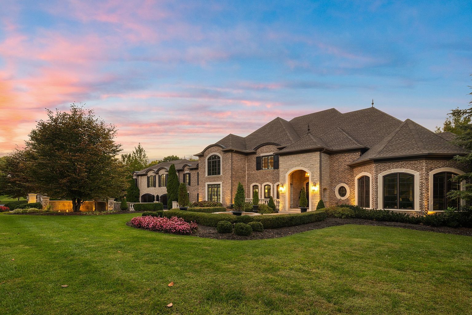 This is a front exterior view of a large, luxurious brick home with a well-manicured lawn and landscaping. The house features multiple arched windows, a prominent entryway with stone accents, and a complex roofline. The overall impression is one of grandeur and sophisticated elegance, enhanced by the soft light of dusk.
