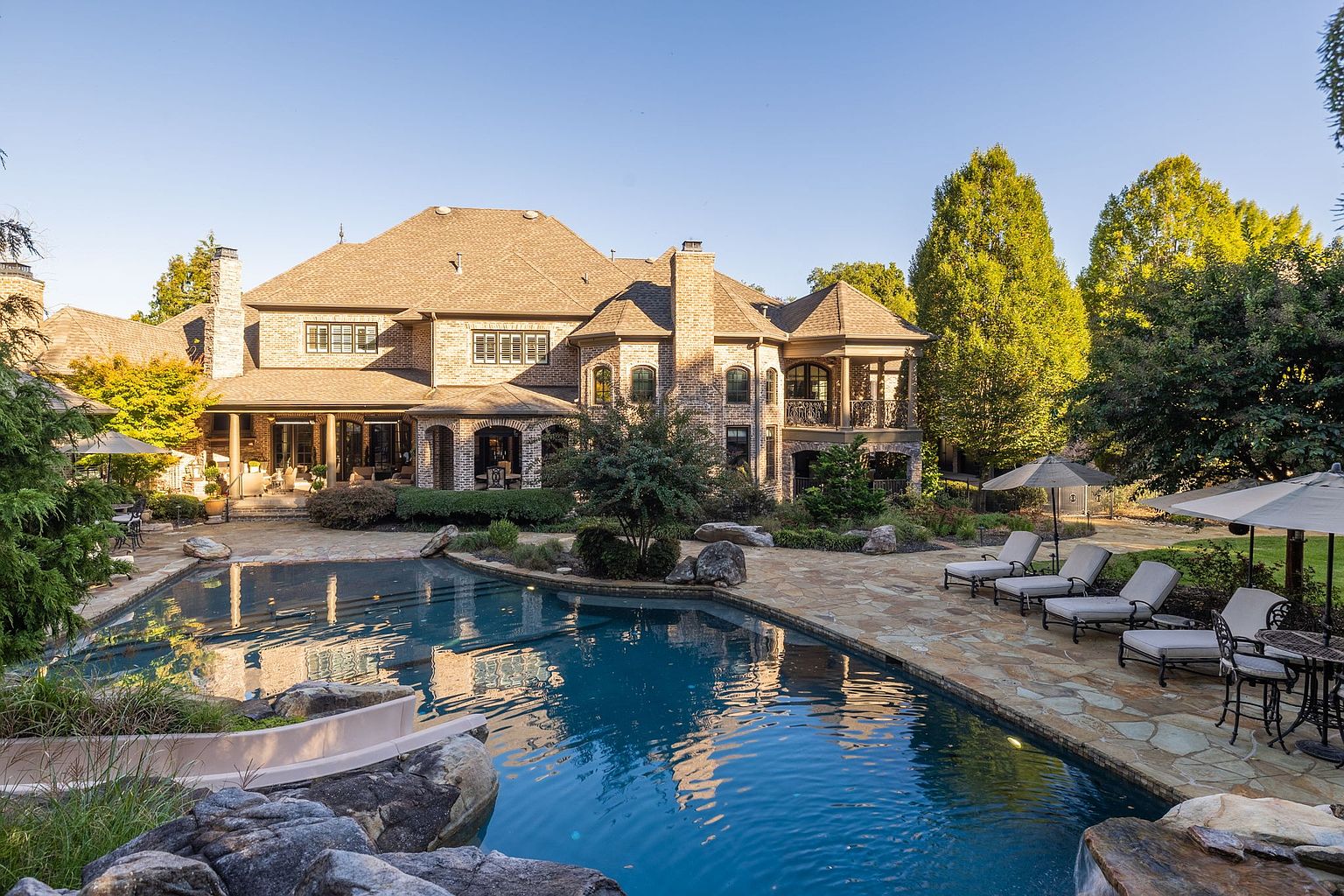 This image showcases a luxurious backyard featuring a large, freeform swimming pool with rock features and a slide. The pool is surrounded by a stone patio with lounge chairs and umbrellas, perfect for relaxation. A grand brick house with multiple stories and balconies serves as a stunning backdrop, creating an inviting and upscale outdoor living space.