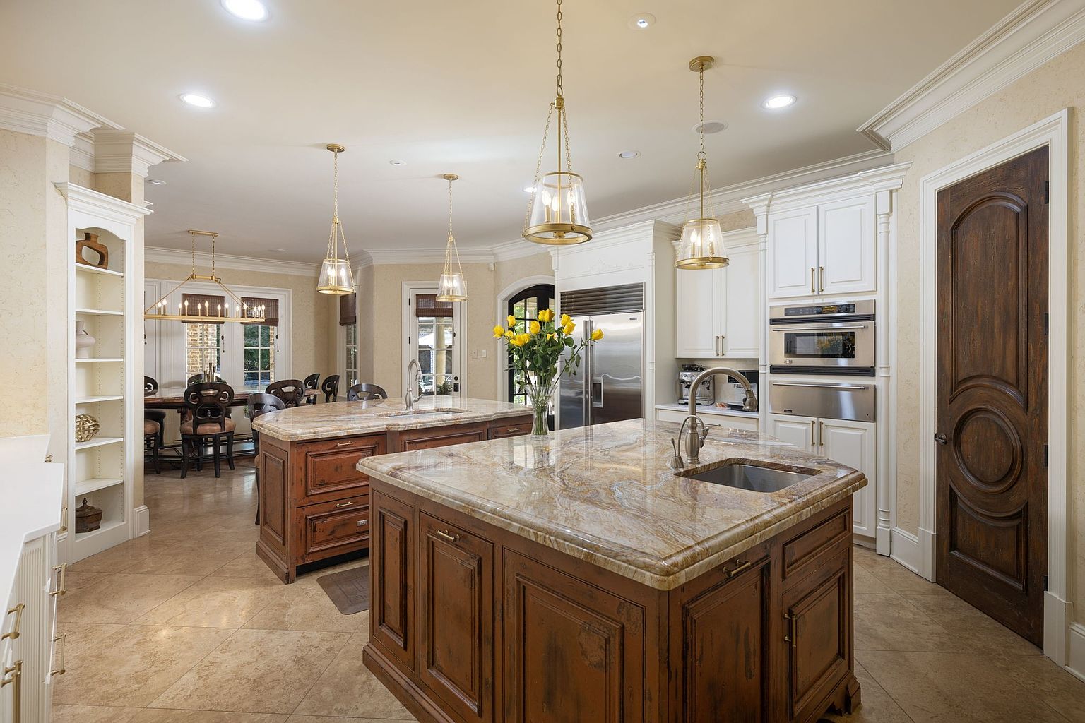 This is a luxurious kitchen featuring two large islands with granite countertops and dark wood cabinetry. The kitchen is equipped with stainless steel appliances and white cabinets along the walls. Pendant lights hang above the islands, and the flooring is a light-colored tile, creating a warm and inviting atmosphere.
