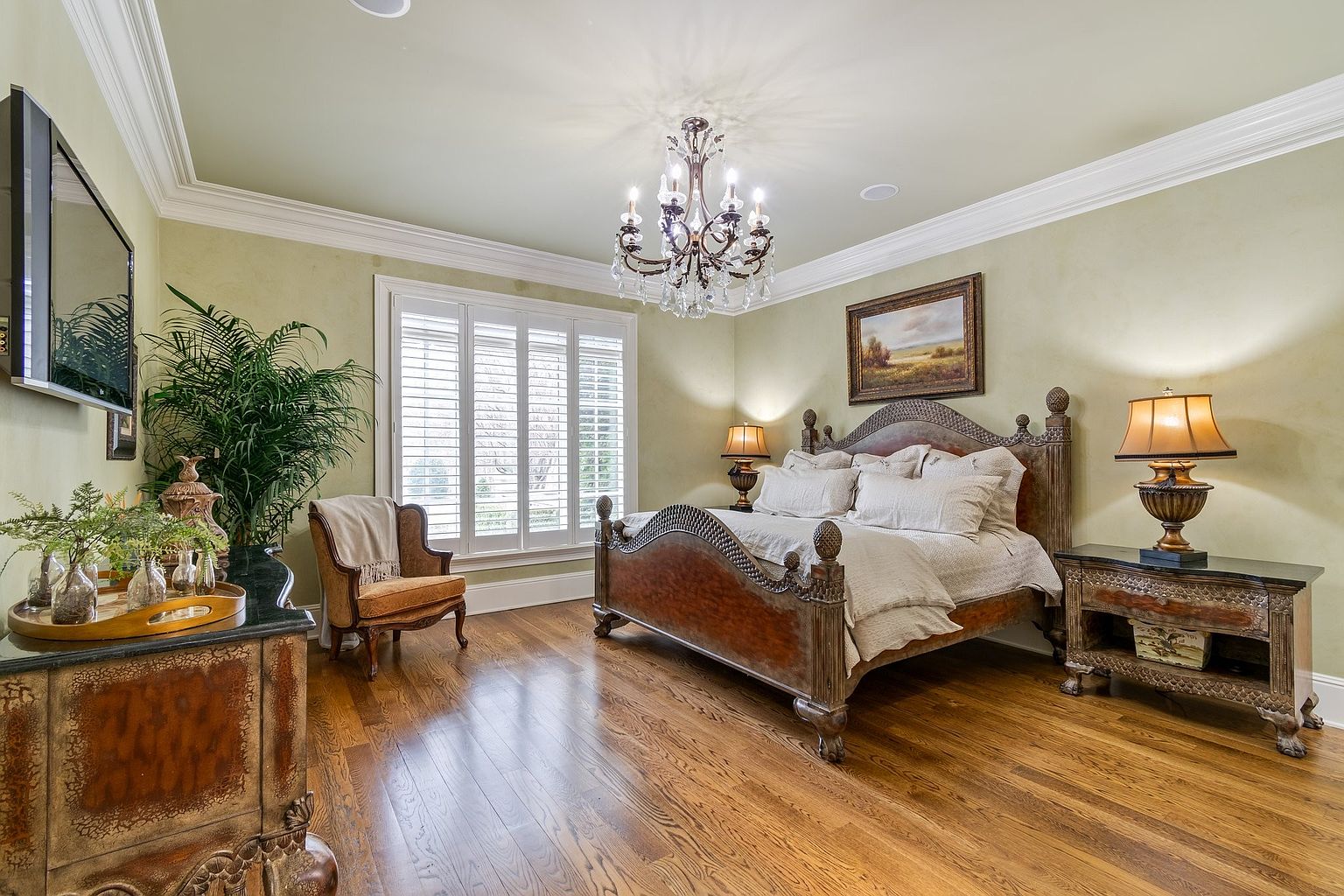 This is a primary bedroom featuring a large, ornate wooden bed with white linens, flanked by matching nightstands and lamps. A crystal chandelier hangs from the ceiling, and a landscape painting adorns the wall above the bed. The room has a traditional style with hardwood floors and light green walls, creating a warm and inviting atmosphere.