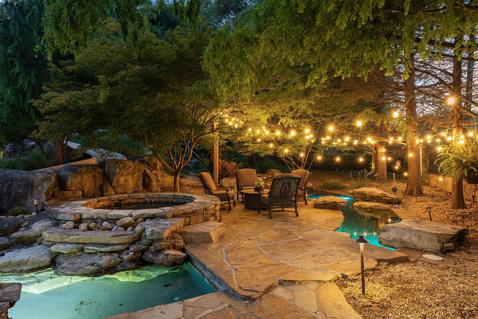 This image showcases a beautifully landscaped backyard featuring a stone patio, a hot tub surrounded by rocks, and a stream leading to a pool. String lights are hung between the trees, creating a warm and inviting ambiance. The scene suggests a luxurious and relaxing outdoor living space, perfect for entertaining or unwinding.