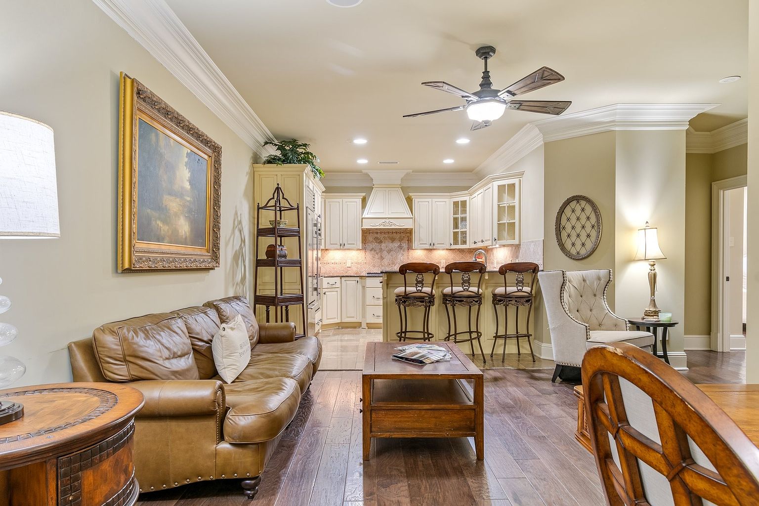 This interior shot showcases a combined living room and kitchen area. The living room features a leather sofa, a framed painting, and a wooden coffee table, while the kitchen boasts white cabinetry, bar stools, and a decorative backsplash. The hardwood flooring and warm lighting create a cozy and inviting atmosphere.