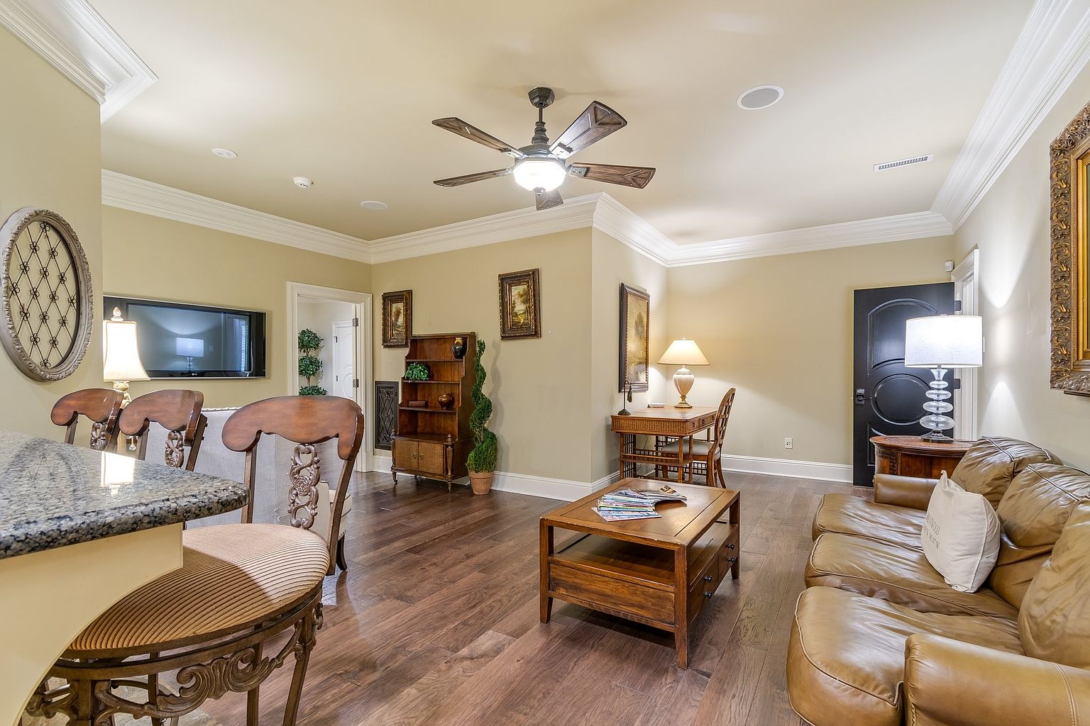 This is an interior shot of a living room featuring a leather sofa, wooden coffee table, and a built-in bookshelf. The room is well-lit with a ceiling fan and lamps, creating a warm and inviting atmosphere. The hardwood flooring and neutral wall colors contribute to a classic and comfortable style.