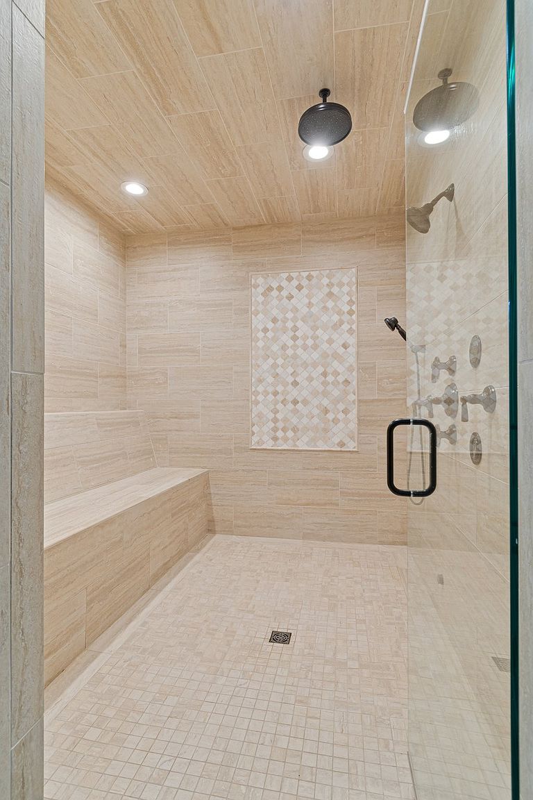 This is a well-lit primary bathroom featuring a spacious walk-in shower with a glass enclosure. The shower is tiled in a neutral, travertine-like material, with a decorative mosaic tile accent wall. A built-in bench provides seating, and a modern shower head and fixtures add to the luxurious feel.