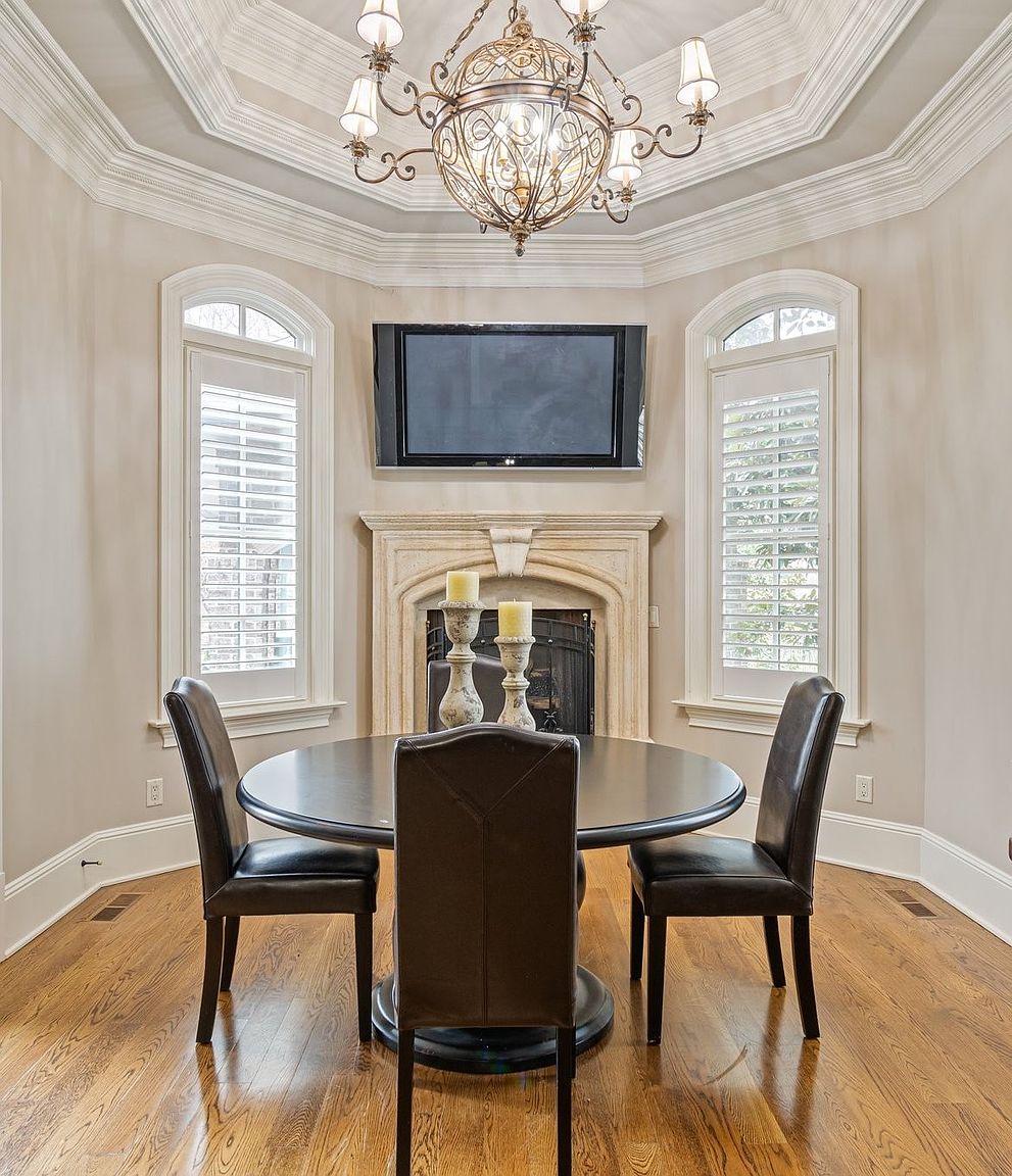This is an interior shot of a dining room featuring a round dark wood table with three leather chairs. A fireplace with a television mounted above it is centered between two arched windows with white shutters. An ornate chandelier hangs from the tray ceiling, illuminating the space and adding a touch of elegance.