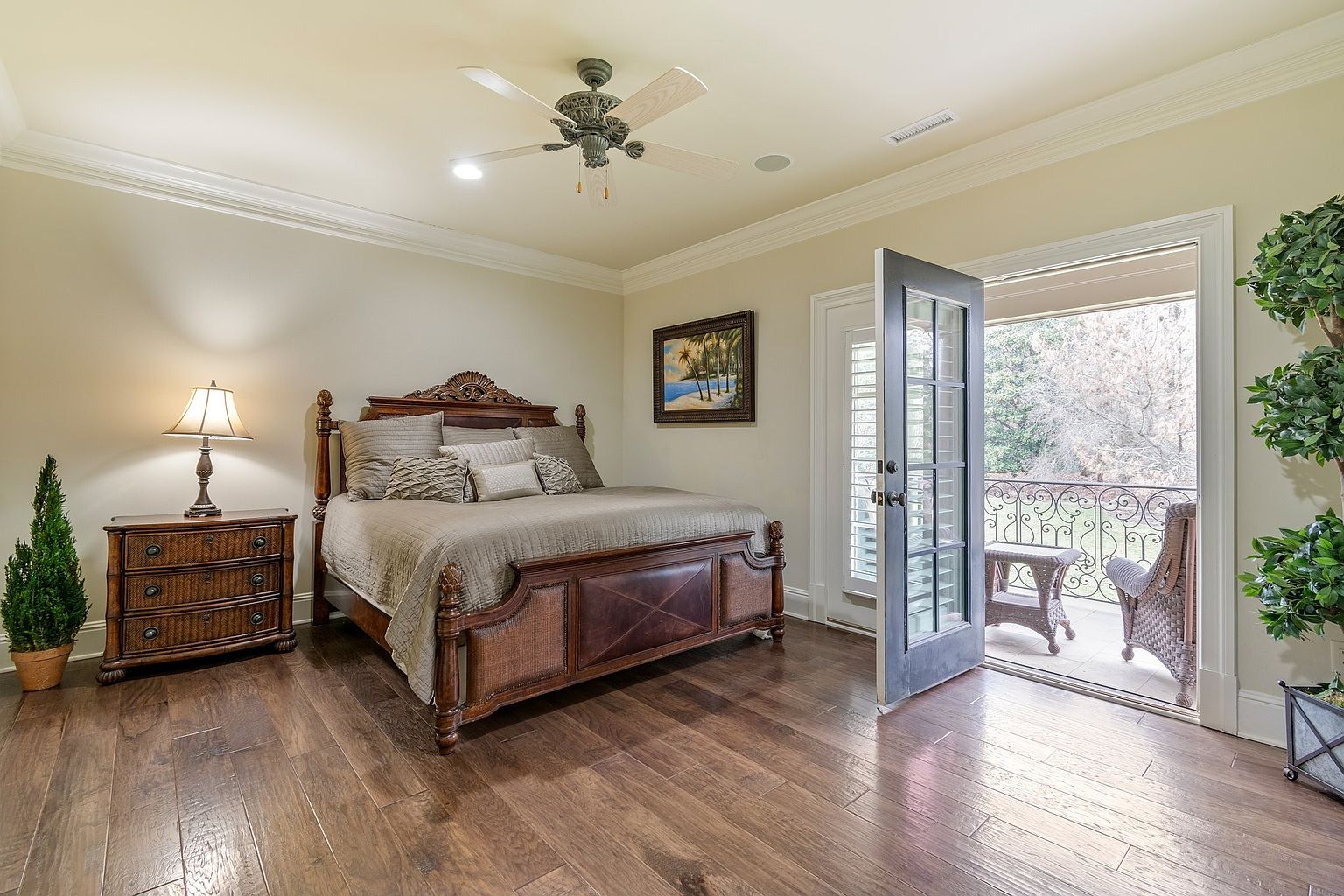 This is a primary bedroom featuring a large, ornate wooden bed with neutral-toned bedding. A matching wooden nightstand with a lamp sits to the left of the bed, and a painting hangs on the wall above the bed. An open door leads to a balcony with wrought iron railings and wicker furniture, creating a seamless indoor-outdoor flow. The room has hardwood floors and a ceiling fan, contributing to a luxurious and comfortable atmosphere.