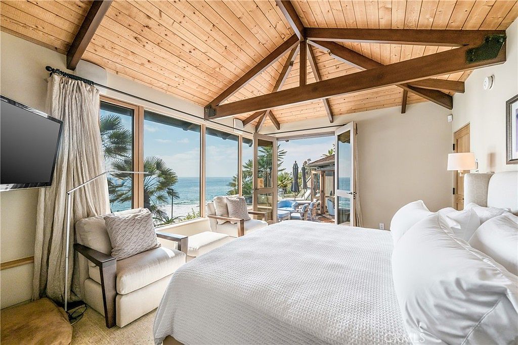 This primary bedroom features a stunning ocean view through large windows and a set of French doors leading to an outdoor patio. The room has a vaulted wood ceiling with exposed beams, creating a warm and inviting atmosphere. The bed is neatly made with white linens, and there are two comfortable armchairs positioned to take advantage of the view.