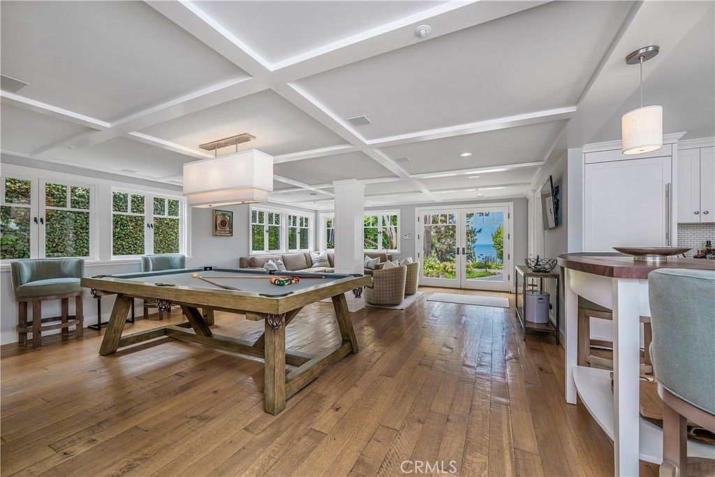 This is a spacious living room featuring hardwood floors, a coffered ceiling, and large windows that provide ample natural light. A pool table is prominently featured, along with comfortable seating arrangements and a view of the outdoors. The room exudes a sense of luxury and relaxation.
