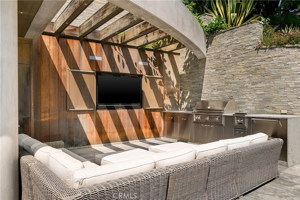 This image showcases a luxurious outdoor living space, featuring a comfortable wicker sectional sofa with light cushions, a built-in stainless steel grill and cabinetry, and a mounted television. The area is covered by a wooden pergola, creating a shaded and inviting atmosphere. The stone wall and lush greenery in the background add to the upscale ambiance, perfect for entertaining or relaxing.