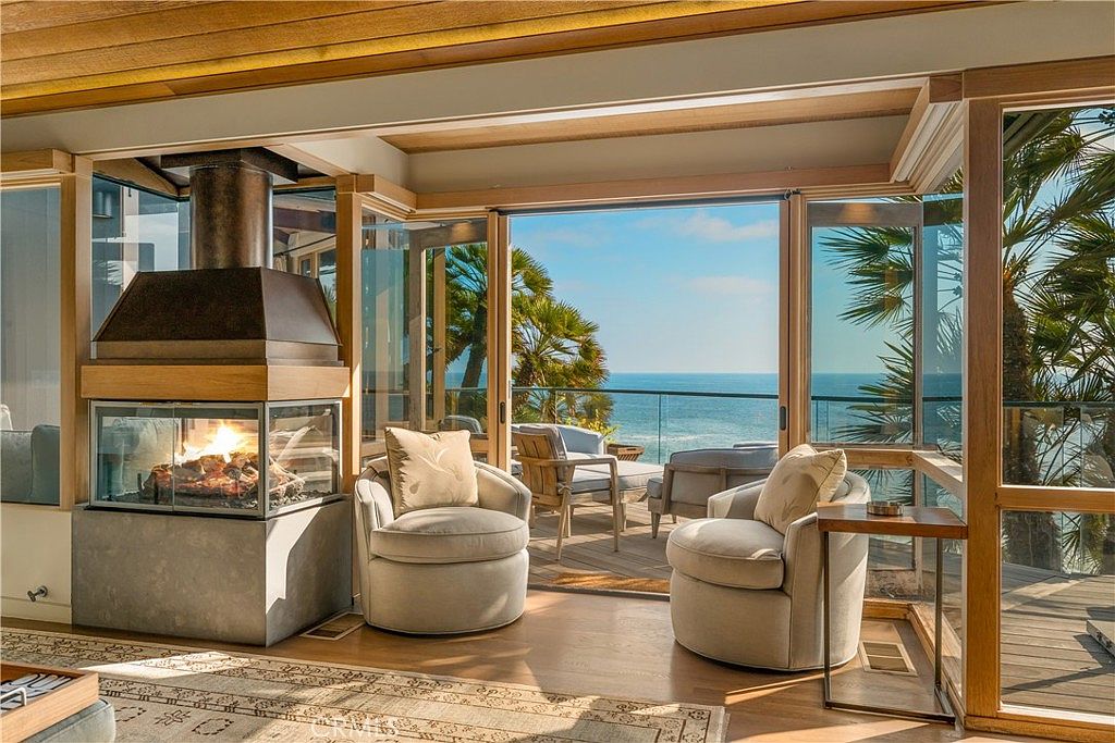 This inviting living room features a modern fireplace, comfortable swivel chairs, and expansive glass doors that open to a stunning ocean view. The room is bathed in natural light, highlighting the warm wood tones and creating a serene and luxurious atmosphere. The open layout seamlessly connects the interior space with the outdoor deck, perfect for enjoying the coastal scenery.