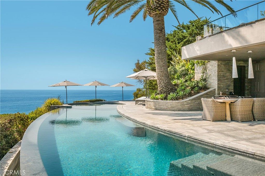 This image showcases a luxurious infinity pool overlooking the ocean. The pool is surrounded by a well-manicured patio area with outdoor seating and a palm tree, creating a serene and upscale ambiance. The architectural design of the adjacent building complements the natural beauty of the coastal setting.