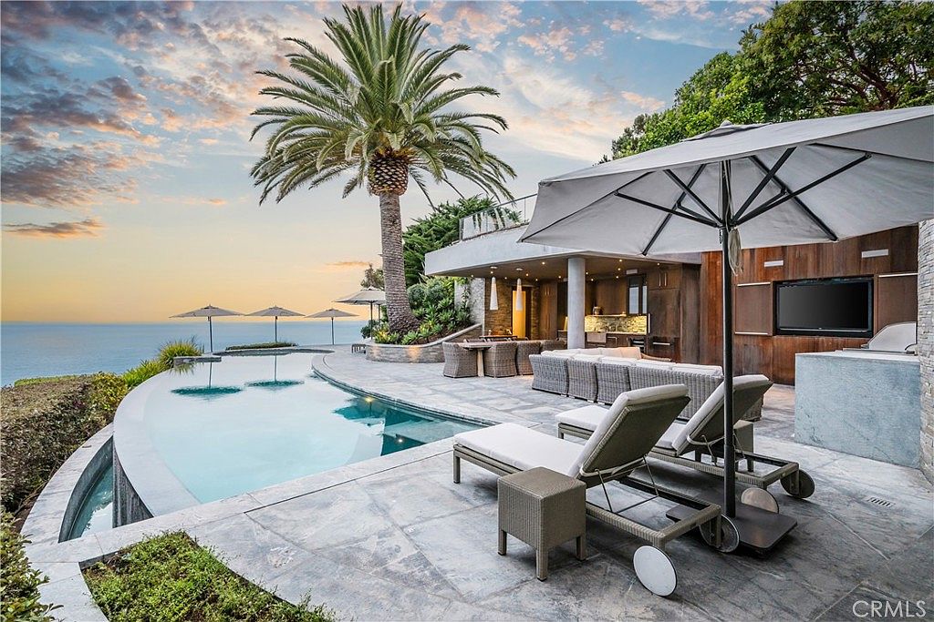 This exterior shot showcases a luxurious pool and spa area with ocean views. The infinity pool blends seamlessly with the horizon, while lounge chairs and an outdoor seating area offer relaxation and entertainment options. A large palm tree adds a tropical touch, enhancing the property's appeal.