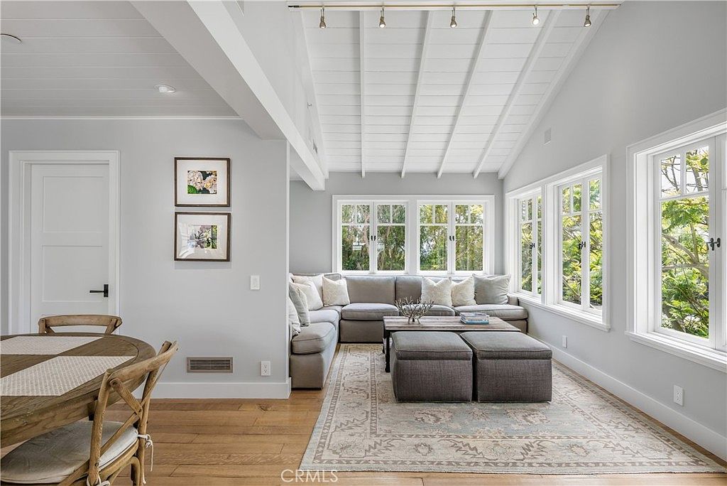 This is an interior shot of a living room featuring a sectional sofa, a coffee table with ottomans, and a large area rug. The room is well-lit with natural light from multiple windows, and the ceiling has a unique design with exposed beams and track lighting. The overall style is modern and comfortable, creating an inviting space.