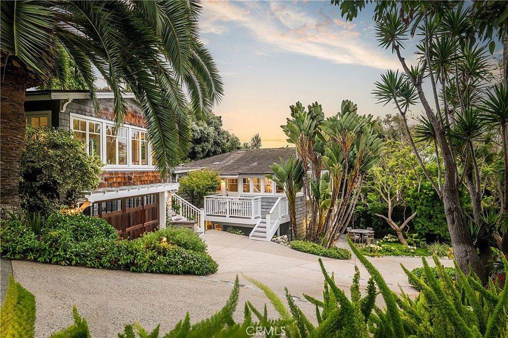 This image showcases the front exterior of a charming home with a multi-level design. The property features a combination of wood siding and shingle details, complemented by lush landscaping including mature palm trees and verdant bushes. A driveway leads up to the house, highlighting the inviting entryway and the overall curb appeal of the residence.