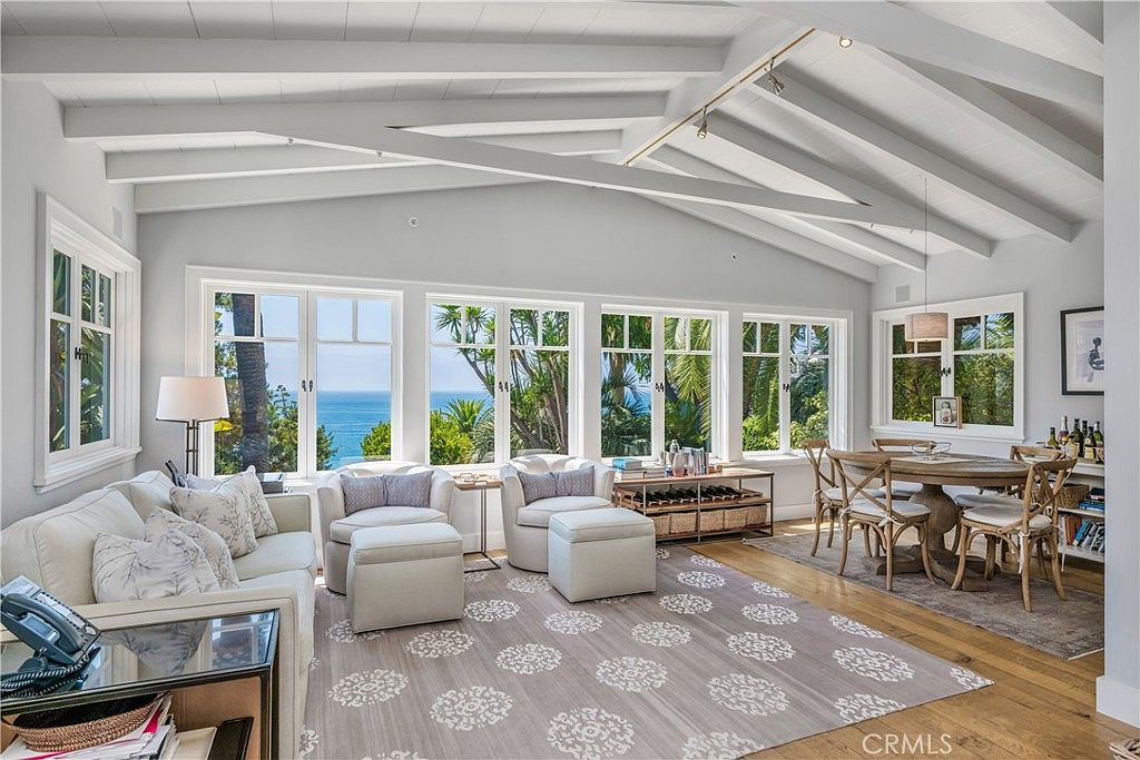 This is an interior shot of a living room with a coastal aesthetic. The room features large windows offering ocean views, a white sofa and armchairs, and a light-colored rug with a floral pattern. The ceiling has exposed beams, adding architectural interest to the space, and a dining area is visible in the background.