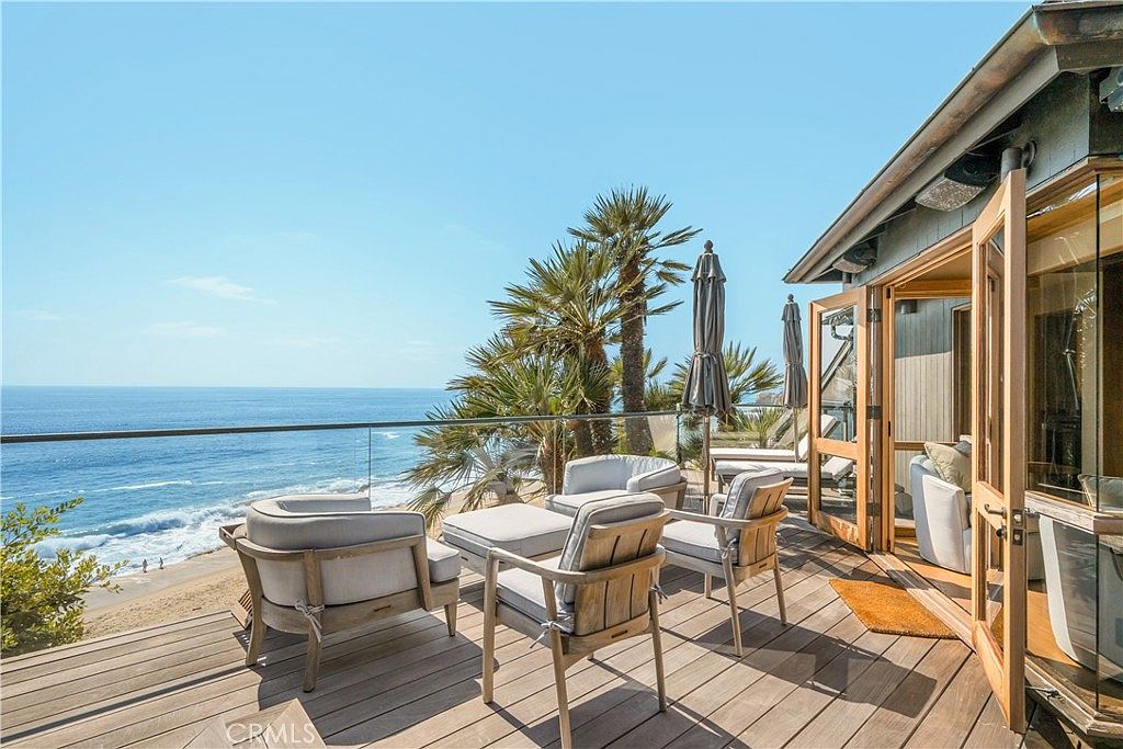 This image showcases a stunning outdoor patio or deck area with breathtaking ocean views. The space is furnished with modern, comfortable seating arrangements, including chairs and a table, perfect for relaxation and entertainment. The deck features a glass railing to maximize the view, and the open doors lead into the home, creating a seamless indoor-outdoor living experience.
