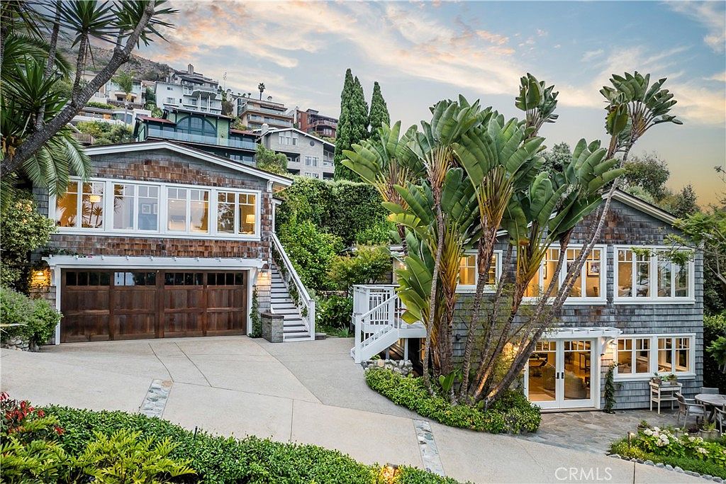 This is a front view of a charming two-story house with a rustic, coastal aesthetic. The exterior features weathered wood shingles, white-framed windows, and a wooden garage door. Lush greenery and mature trees surround the property, adding to its curb appeal and creating a sense of privacy.