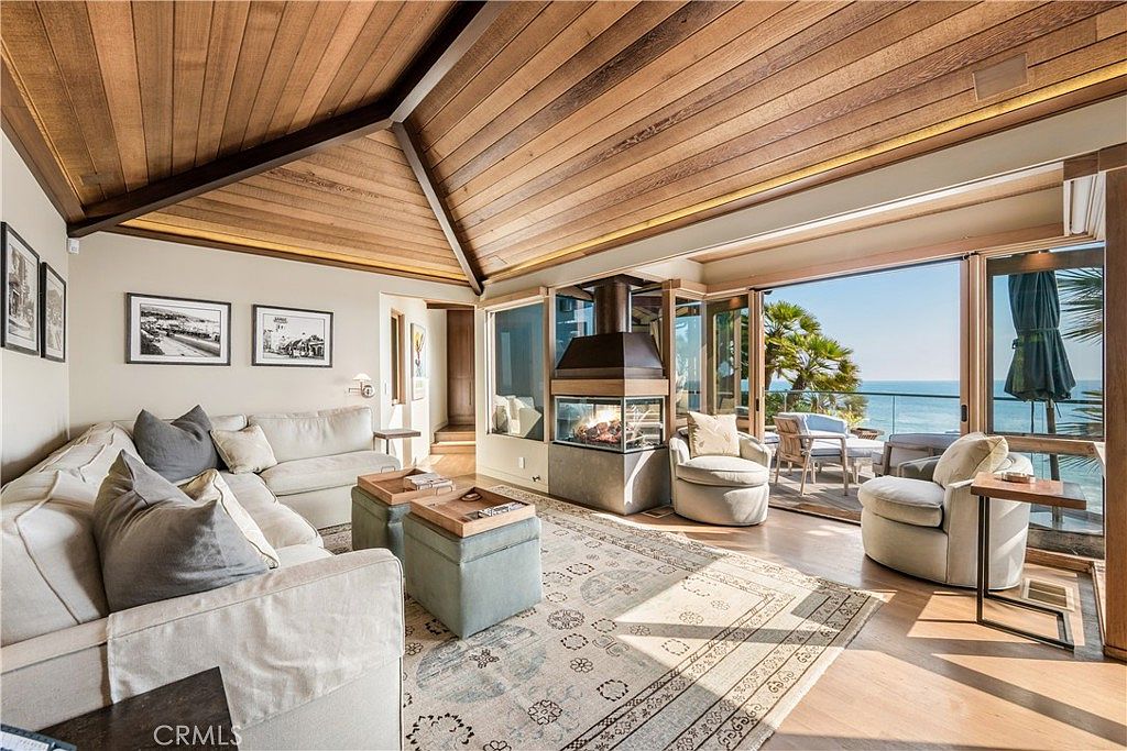 This is a bright and airy living room featuring a modern fireplace, a comfortable sectional sofa with throw pillows, and a large area rug. The room has a high, wood-paneled ceiling and large glass doors that open to a patio with ocean views. The overall style is luxurious and inviting, perfect for relaxation and entertaining.