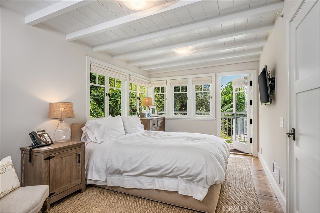 This is a bright and airy primary bedroom featuring a large bed with white linens, a wooden nightstand with a lamp, and multiple windows offering natural light and views of greenery. The room has a neutral color palette, white beamed ceilings, and a door leading to a balcony, creating a serene and inviting atmosphere. A television is mounted on the wall.