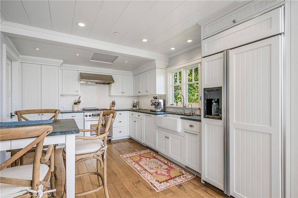 This is a bright and airy kitchen featuring white cabinetry, a farmhouse sink, and stainless steel appliances. The kitchen has a classic, clean aesthetic with a wooden floor and a patterned rug adding warmth. A small dining table with chairs is visible in the foreground, suggesting a cozy breakfast nook.