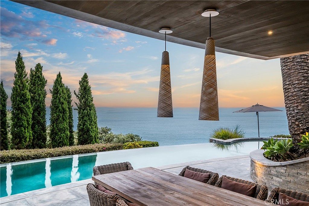 This image showcases a luxurious outdoor patio/deck area with stunning ocean views. The space features a wooden dining table with wicker chairs, pendant lighting, and an infinity pool that blends seamlessly with the horizon. Lush greenery and palm trees add to the tropical ambiance, creating an inviting and upscale outdoor living space.