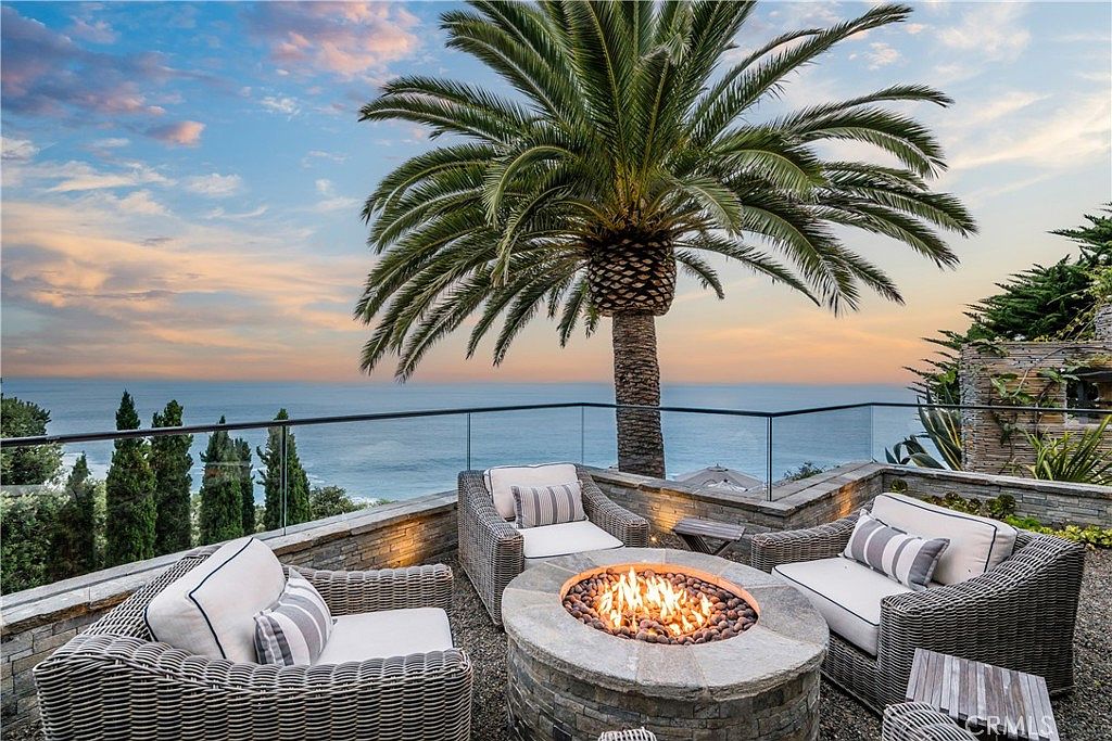 This image showcases a luxurious outdoor patio or deck area with a stunning ocean view. The space is furnished with comfortable wicker seating arranged around a stone fire pit, creating an inviting atmosphere. A tall palm tree adds a tropical touch, while a glass railing ensures unobstructed views of the ocean and sky, perfect for relaxation and entertainment.