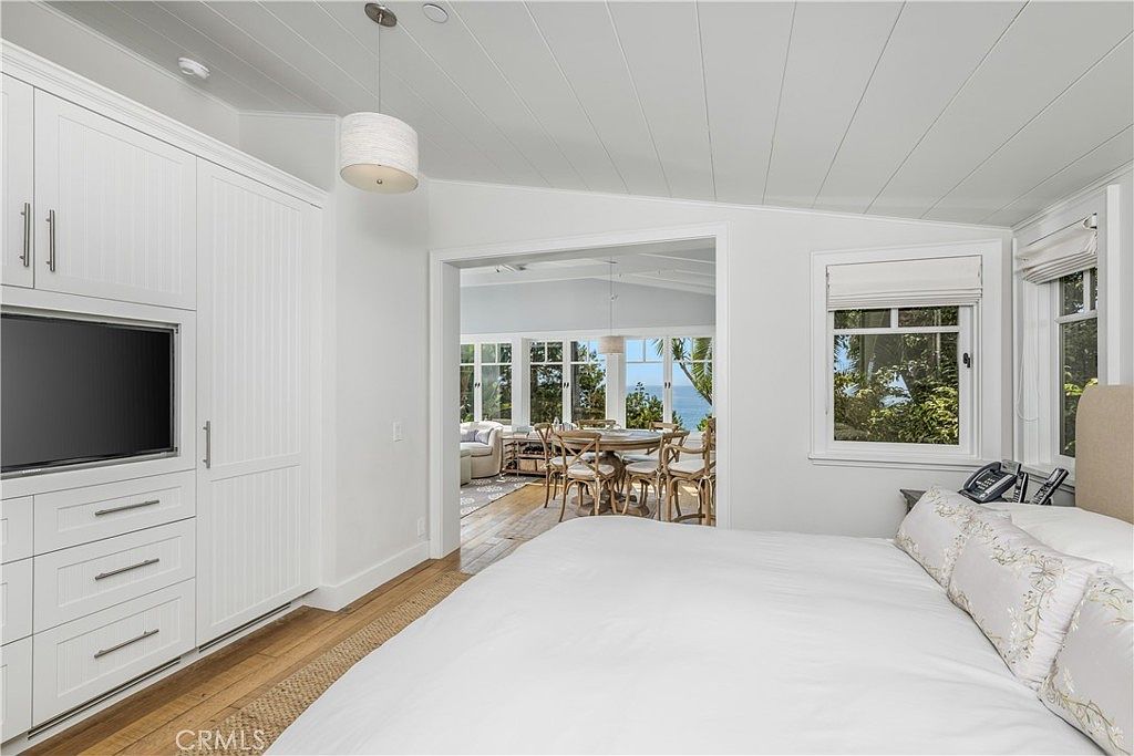 This is a bright and airy primary bedroom featuring a large bed with white linens and decorative pillows. The room has built-in white cabinetry with a television, and a doorway leads to a sunroom or dining area with views of the ocean. A window provides natural light, and the overall style is coastal and serene.