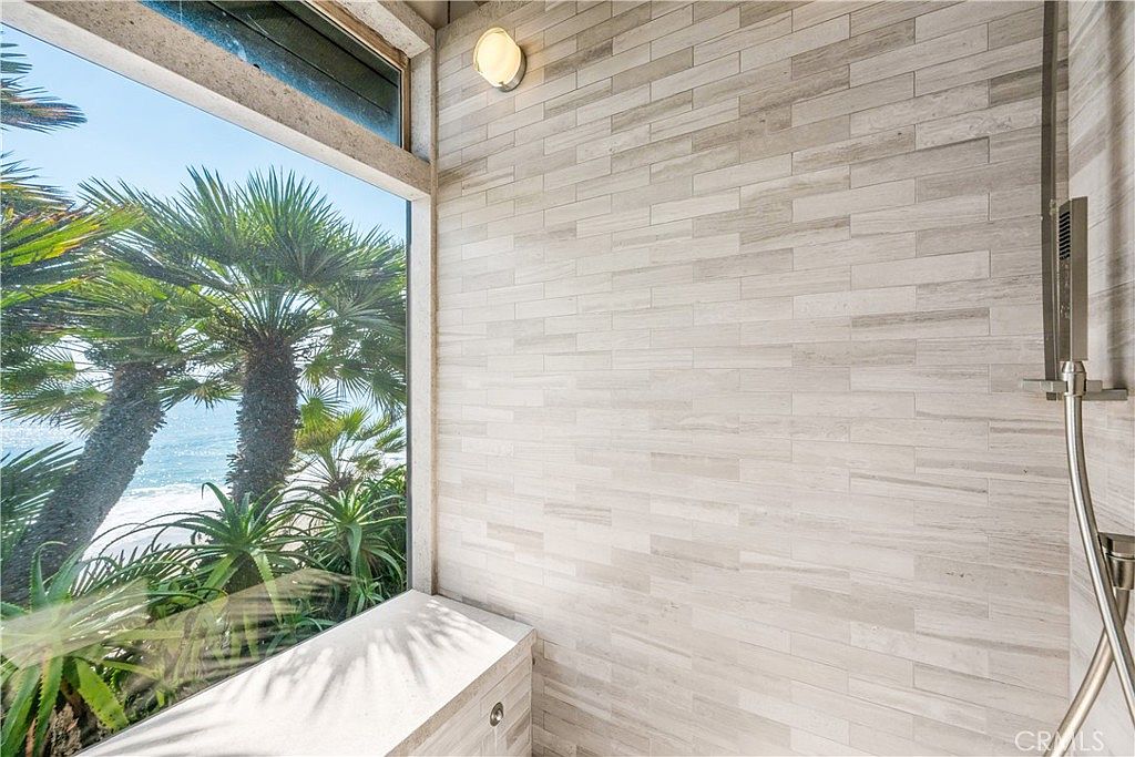 This is a bright and airy primary bathroom featuring a walk-in shower with a large window overlooking lush greenery and the ocean. The shower is tiled with light-colored, horizontally laid rectangular tiles, and a modern shower fixture is visible. A built-in bench sits beneath the window, enhancing the spa-like atmosphere.