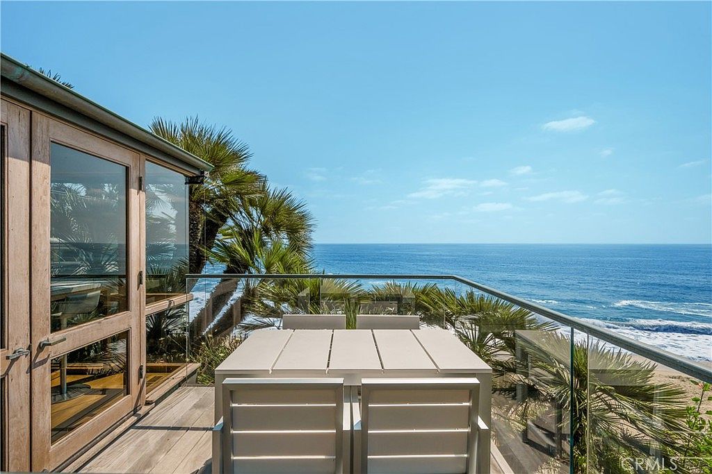 This image showcases a beautiful outdoor patio or balcony with stunning ocean views. The space features a modern dining set with a white table and chairs, set against a backdrop of palm trees and a clear glass railing. The overall impression is one of luxury and relaxation, perfect for enjoying the coastal scenery.