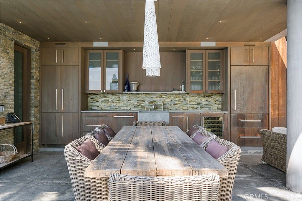 This image showcases an elegant outdoor dining area on a patio or deck. The space features a large wooden dining table surrounded by wicker chairs with plush cushions. Built-in cabinetry with a sink and backsplash provides a functional outdoor kitchen area, enhancing the space for entertaining and relaxation.