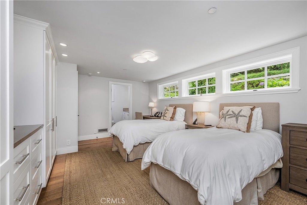 This is a bright and inviting guest bedroom featuring two twin beds with white linens and decorative pillows. The room has a neutral color palette with white walls and light wood flooring, complemented by natural light streaming through the windows. A built-in wardrobe provides ample storage space, enhancing the room's functionality and appeal.