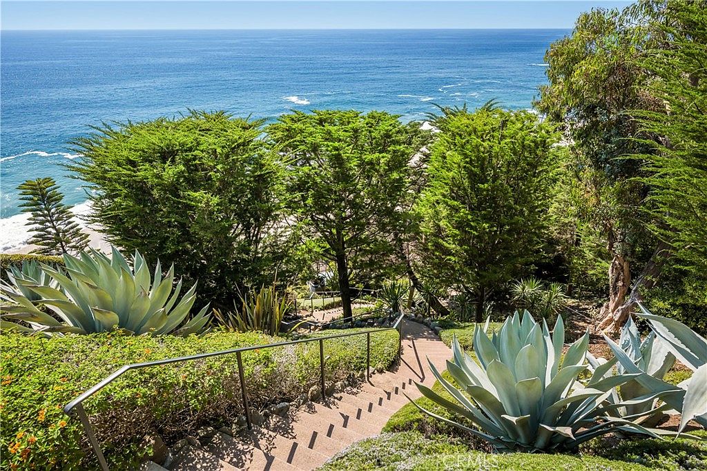This image showcases a lush yard or garden leading down to the ocean. A set of stairs with railings descends through the greenery, which includes agave plants and various shrubs. The view culminates in a stunning ocean vista, creating a serene and inviting atmosphere.