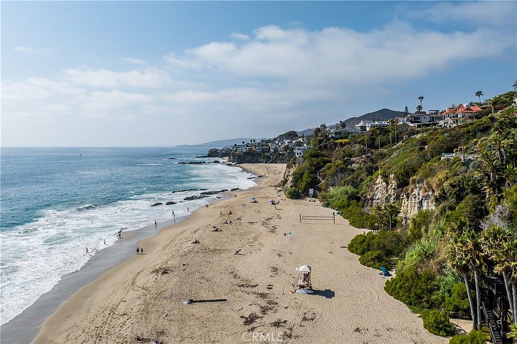 This aerial view showcases a stunning coastal property with a sandy beach, turquoise ocean waters, and lush hillside landscaping. Several houses are nestled on the hillside, offering breathtaking ocean views. The scene is bathed in sunlight, creating a serene and inviting atmosphere, perfect for highlighting the property's prime location and lifestyle appeal.