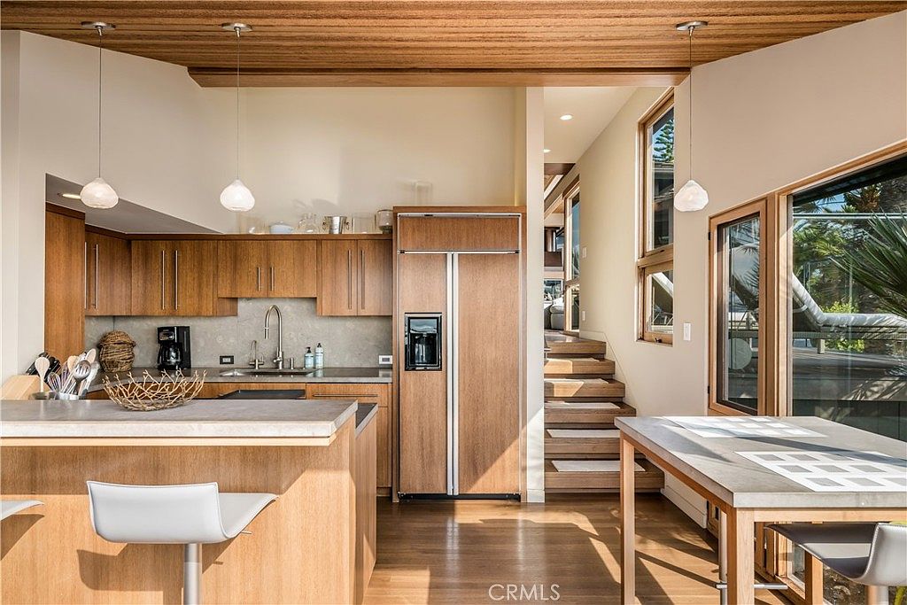 This interior shot showcases a modern kitchen with wooden cabinetry and a contrasting countertop island. The kitchen seamlessly transitions into a dining area with a wooden table and chairs, and a staircase is visible in the background, suggesting an open floor plan. Natural light floods the space through large windows, highlighting the warm tones of the wood and creating an inviting atmosphere.