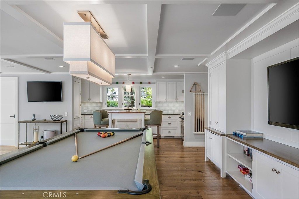 This interior shot showcases a living room area that seamlessly blends entertainment and relaxation. A pool table takes center stage, complemented by a nearby television and custom cabinetry. The hardwood flooring and coffered ceiling add a touch of elegance to the space.