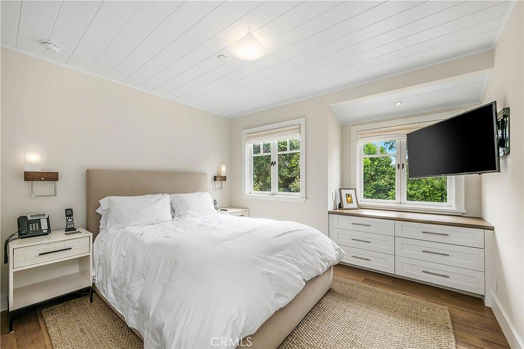This is a bright and inviting primary bedroom featuring a queen-sized bed with white linens and a neutral-toned headboard. The room has a clean, modern aesthetic with white walls, a wood-planked ceiling, and built-in storage beneath the windows. A wall-mounted television and bedside tables with lamps add to the room's functionality and comfort.
