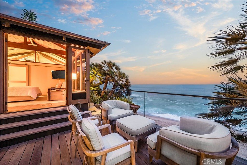 This image showcases a luxurious patio/deck area with comfortable outdoor seating overlooking a stunning ocean view. The deck features wooden flooring and a glass railing, providing an unobstructed view of the beach and ocean. The scene is set at sunset, with warm colors in the sky, creating a serene and inviting atmosphere.
