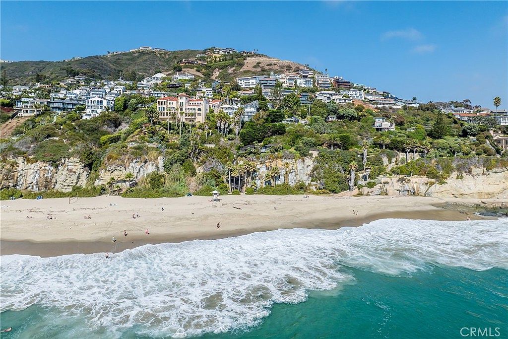 This aerial view showcases a stunning coastal property with a hillside of luxurious homes overlooking a sandy beach and the ocean. The architecture is diverse, with Mediterranean-style villas and modern residences nestled among lush greenery. The turquoise water and crashing waves add to the picturesque setting, creating an idyllic and desirable location.