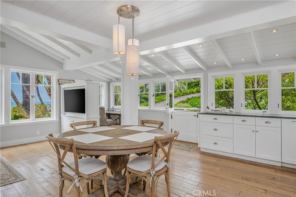 This is an interior shot of a dining room featuring a round wooden table with four chairs. The room has white walls, a white beamed ceiling, and multiple windows offering natural light and views of the outdoors. A modern chandelier hangs above the table, and built-in white cabinets are visible along one wall.