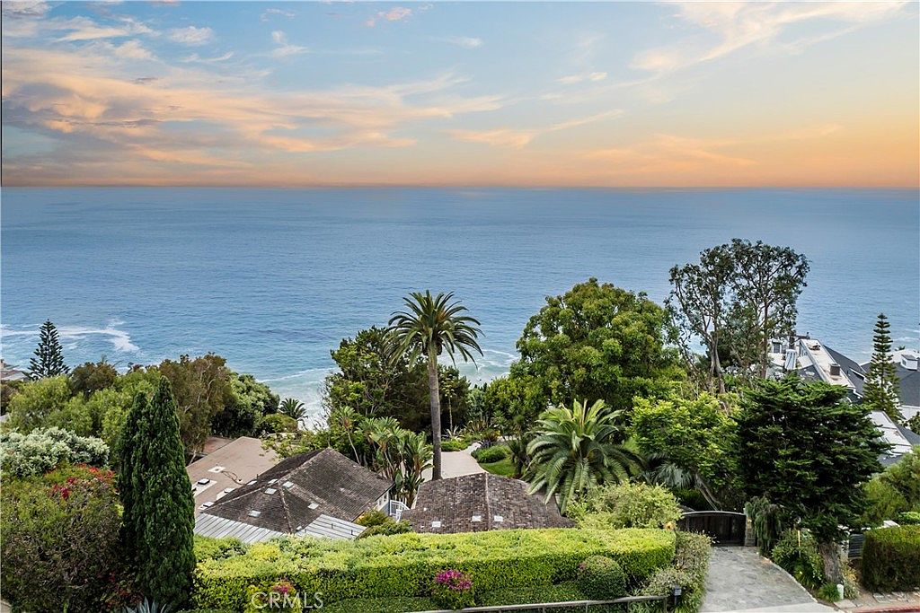 This aerial view showcases a luxurious property with lush greenery, including mature trees and manicured hedges. The rooftops of the houses are visible, suggesting a multi-level design. In the background, the expansive ocean creates a serene and picturesque setting, enhancing the property's appeal.