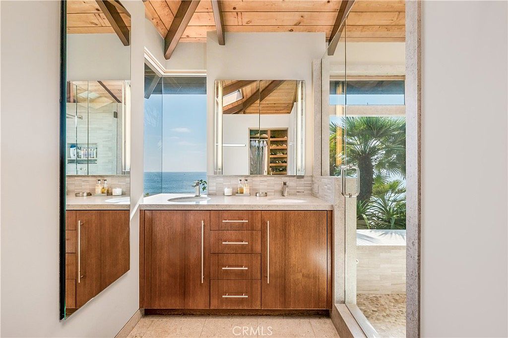 This is a luxurious primary bathroom featuring a double vanity with wood cabinetry and light countertops. Large mirrors reflect the space and the ocean view visible through a window, while a glass-enclosed shower area is visible to the right. The bathroom exudes a spa-like atmosphere with natural light and high-end finishes.