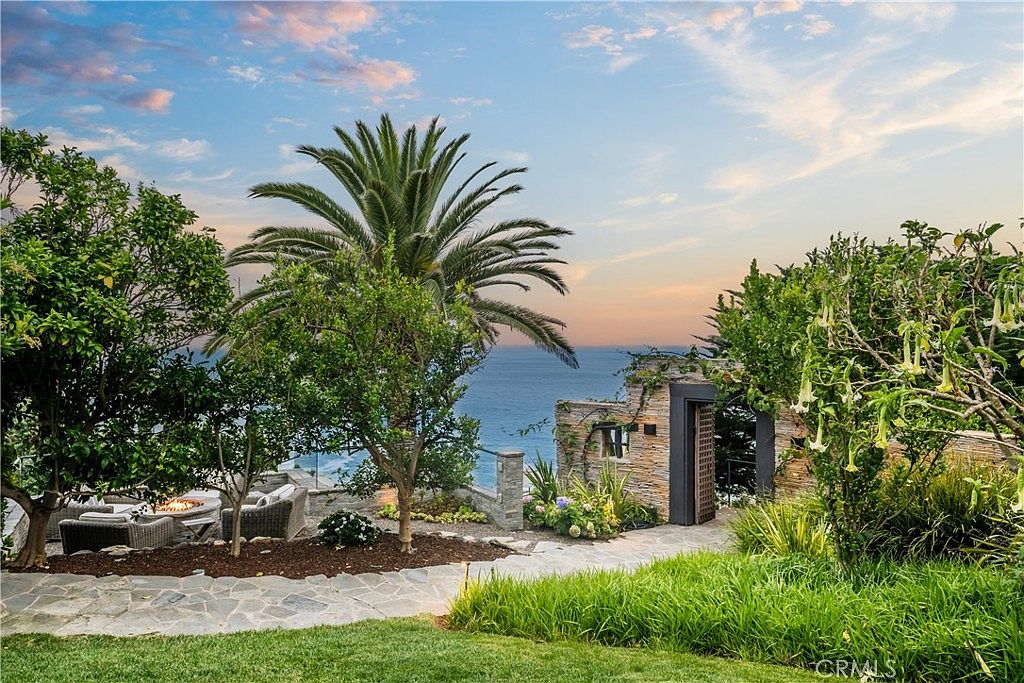 This image showcases a beautifully landscaped yard with a stunning ocean view. A stone pathway leads to a gated entrance, surrounded by lush greenery, including a palm tree and flowering bushes. A cozy seating area with a fire pit is visible in the background, creating an inviting outdoor space.