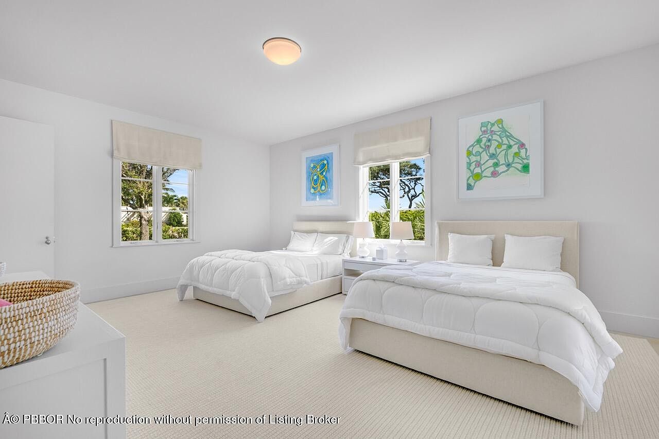 This is a bright and airy guest bedroom featuring two twin beds with white linens and light beige headboards. Natural light floods the room through two windows adorned with neutral-toned roman shades. Artwork hangs above each bed, adding a touch of color to the otherwise white walls and light carpet, creating a serene and inviting space.
