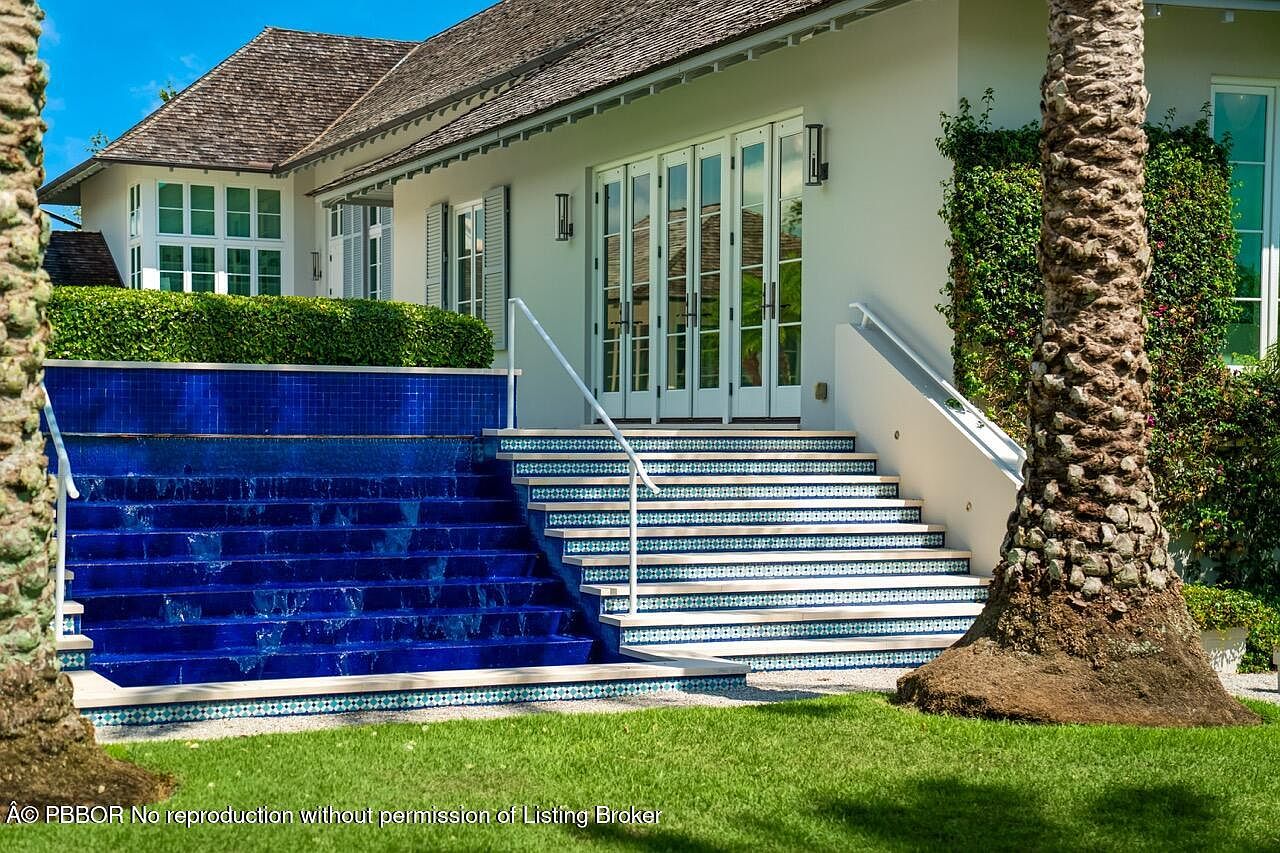 This image showcases the front exterior of a luxurious home, featuring a unique water feature cascading down blue-tiled steps leading to the entrance. The house is painted in a neutral tone, complemented by white trim and multiple sets of French doors. Lush greenery surrounds the property, adding to its curb appeal and creating a serene atmosphere.